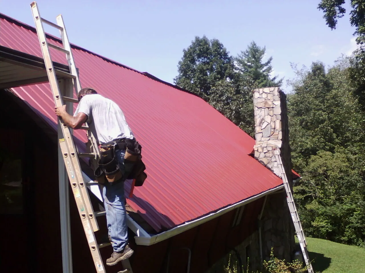 Roofer on a ladder beside a red metal roof and stone chimney — Soffit & Fascia Repair by Roofing Brothers in Fairbury